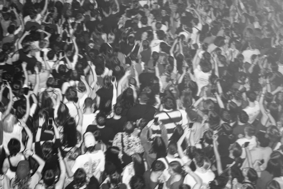 Crowd of people at a large indoor concert or event, many raising their hands.