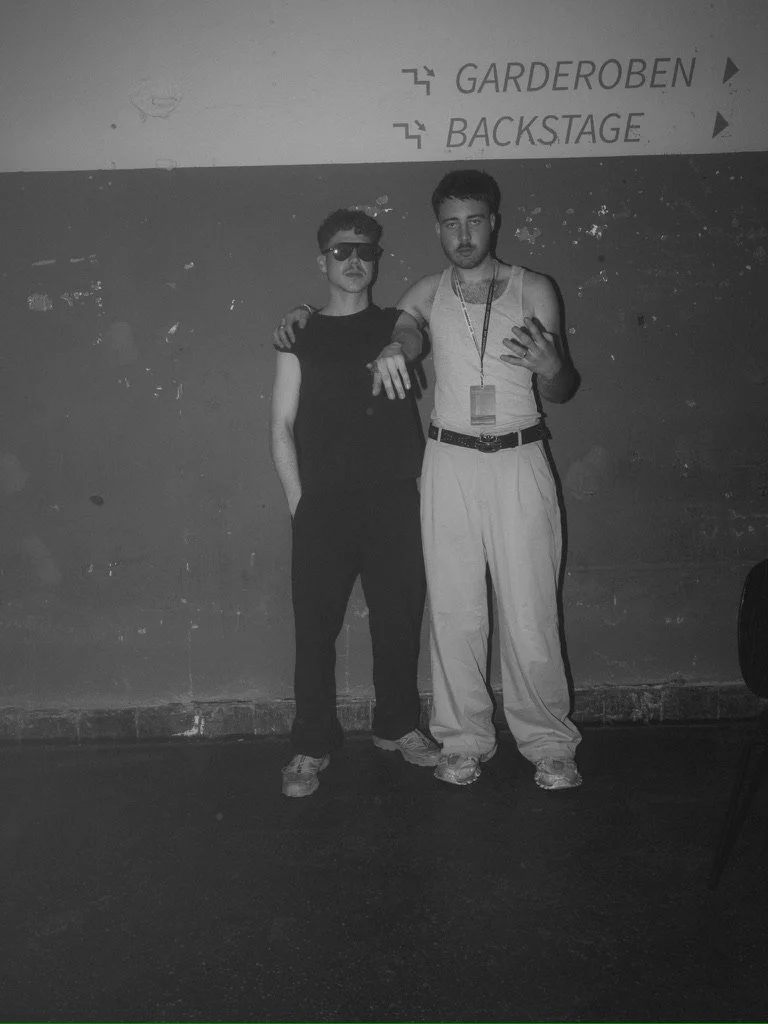 Two young men standing in front of a wall with signs for 'GARDEROBEN' and 'BACKSTAGE', posing for a photo inside a building, one wearing sunglasses and the other with a lanyard around his neck.