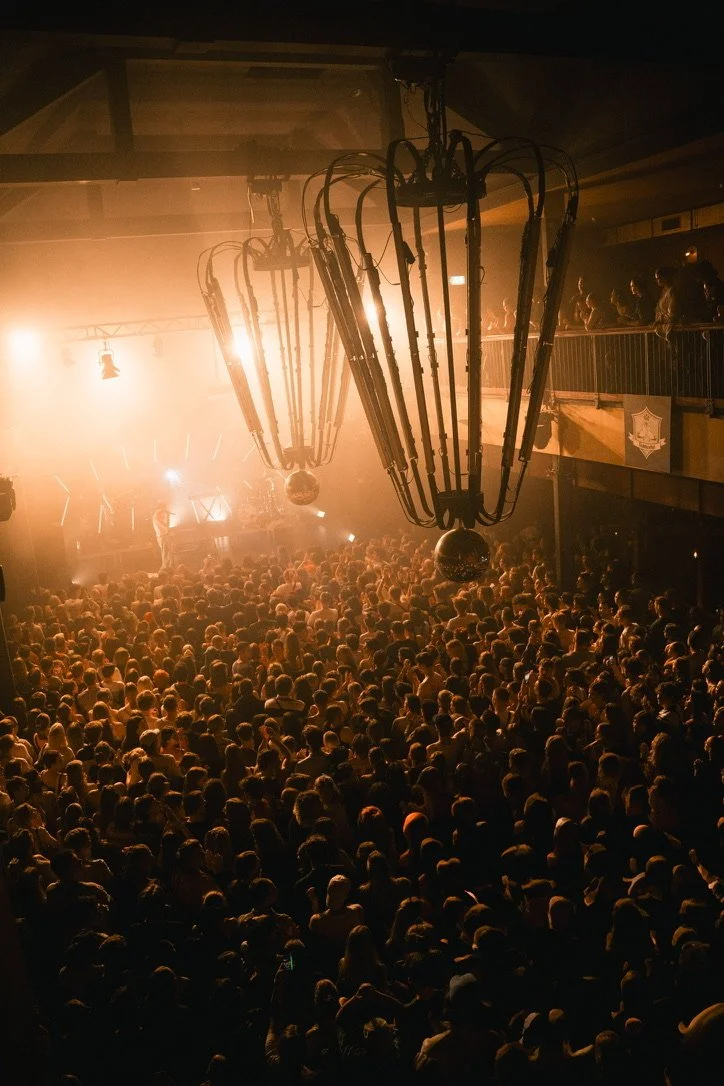 A crowded nightclub with a large number of people dancing and enjoying the event. The space features hanging chandeliers, a DJ booth with bright lights, and a balcony with additional spectators.