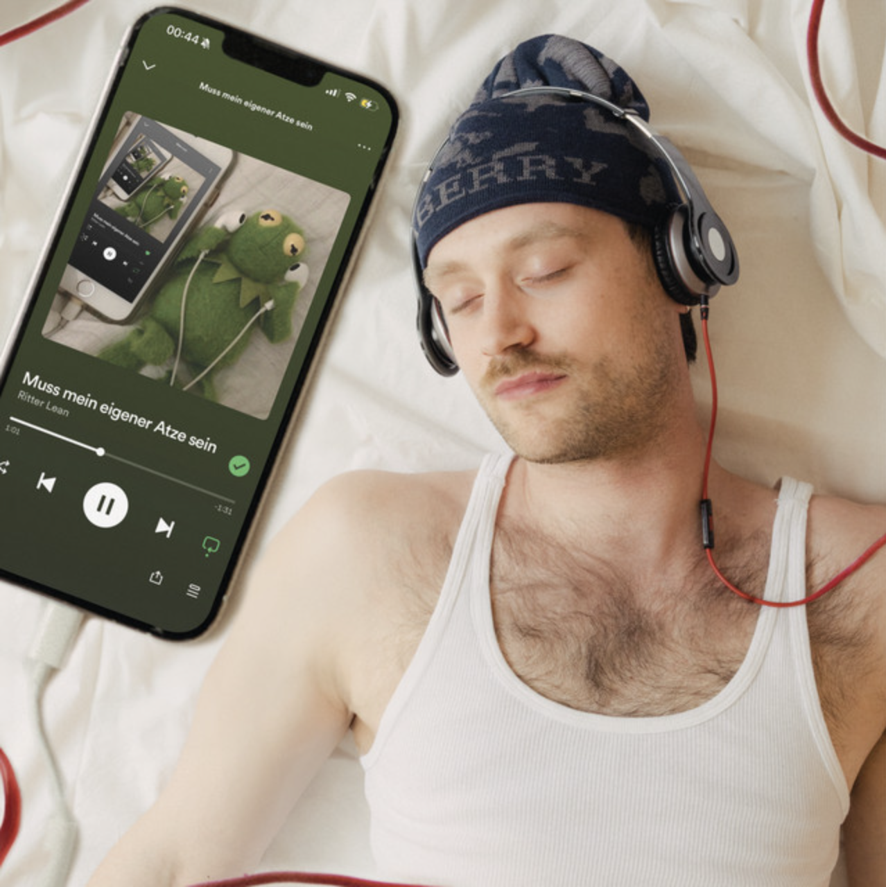 A man sleeping on a bed with headphones on, a tank top, next to a smartphone playing music, showing a picture of a stuffed Kermit the Frog puppet also connected to the phone via headphones.