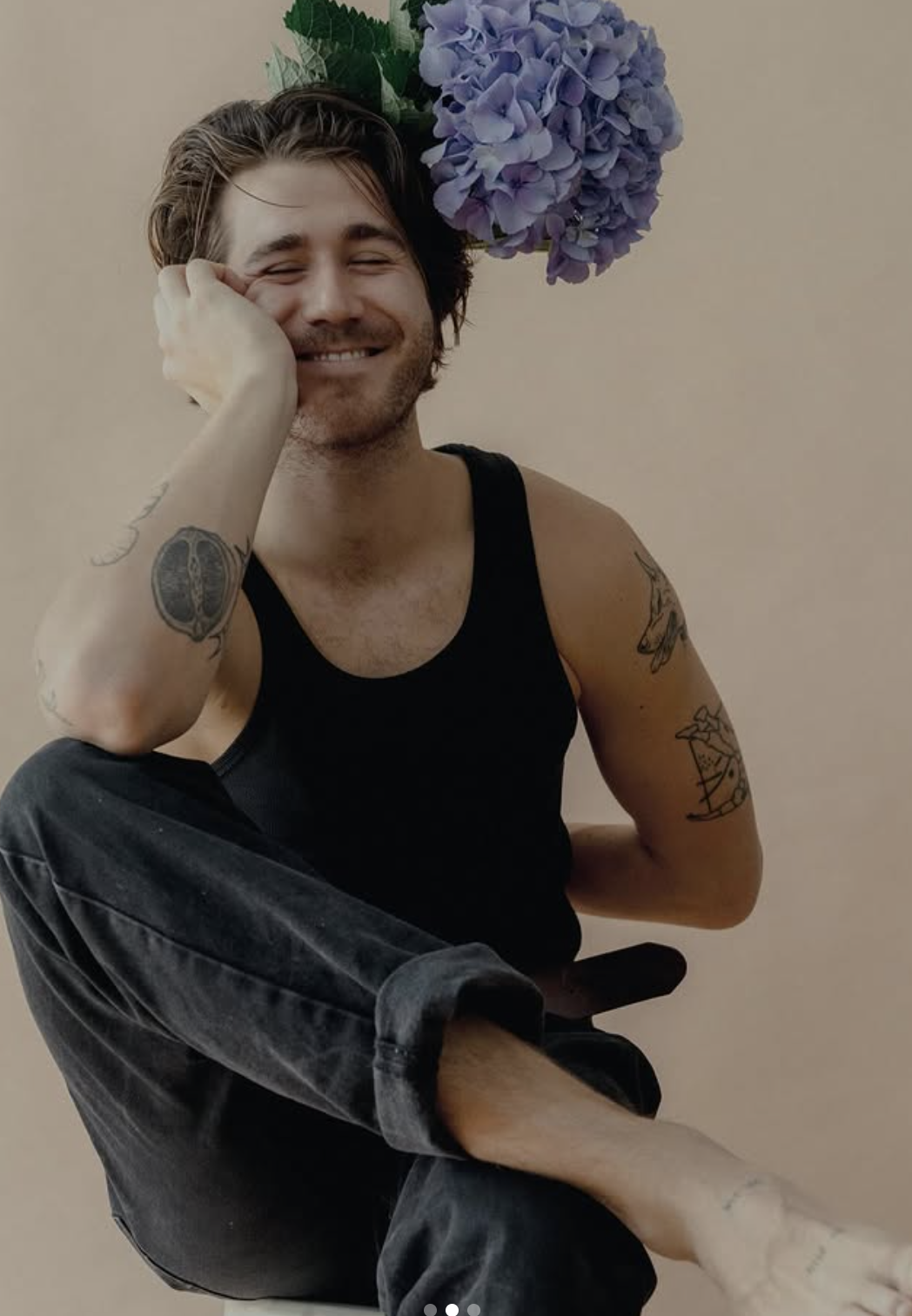 A young man with glasses and tattoos on his arms, wearing a black tank top and black jeans, smiling and sitting with his knee up, with purple hydrangea flowers nearby.