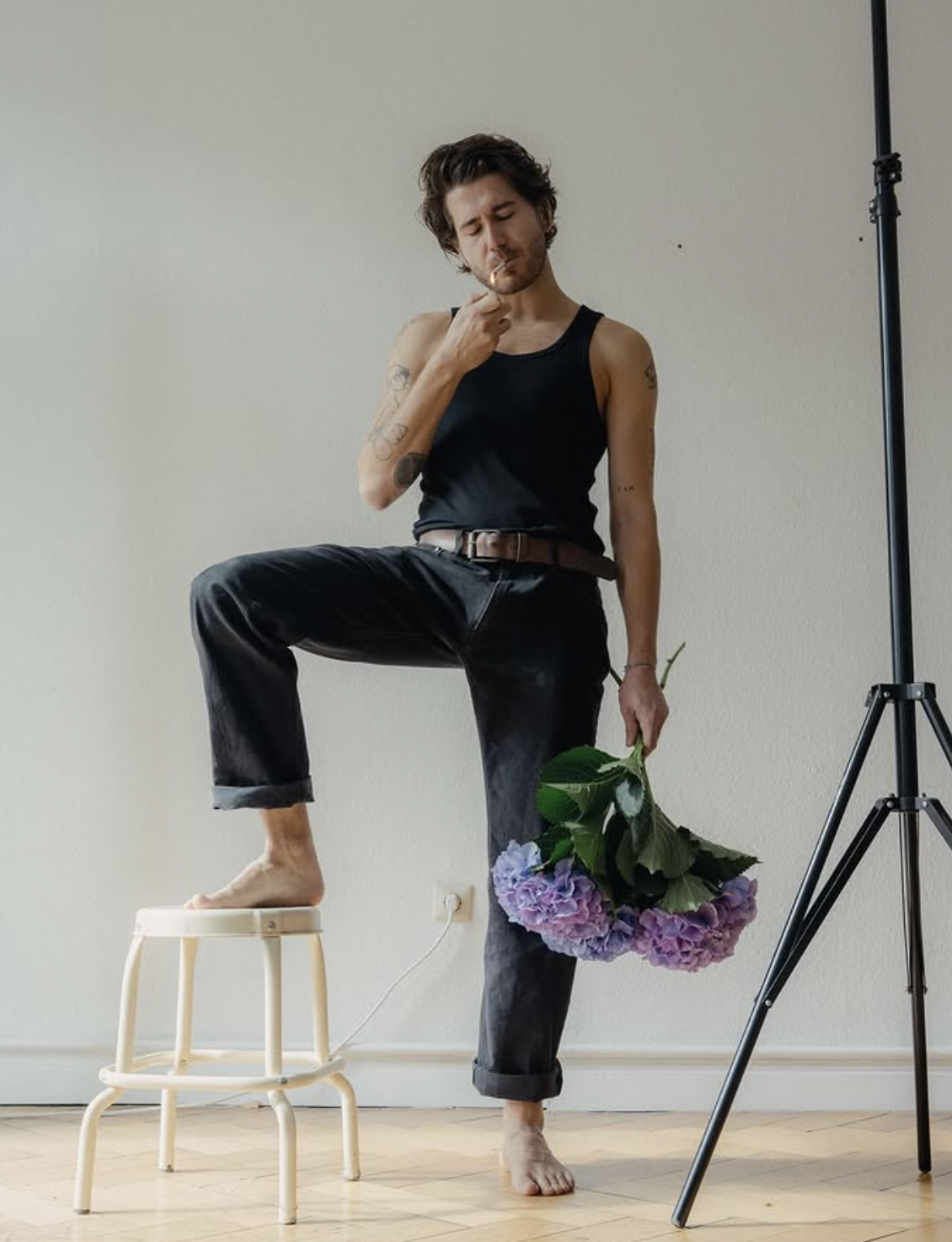 A young man with dark hair and tattoos, dressed in a black tank top and jeans, stands with one foot on a small wooden stool while holding a bouquet of purple hydrangeas. He appears to be in a studio with a plain off-white wall, a tripod stand on the right, and a power outlet on the wall behind him.