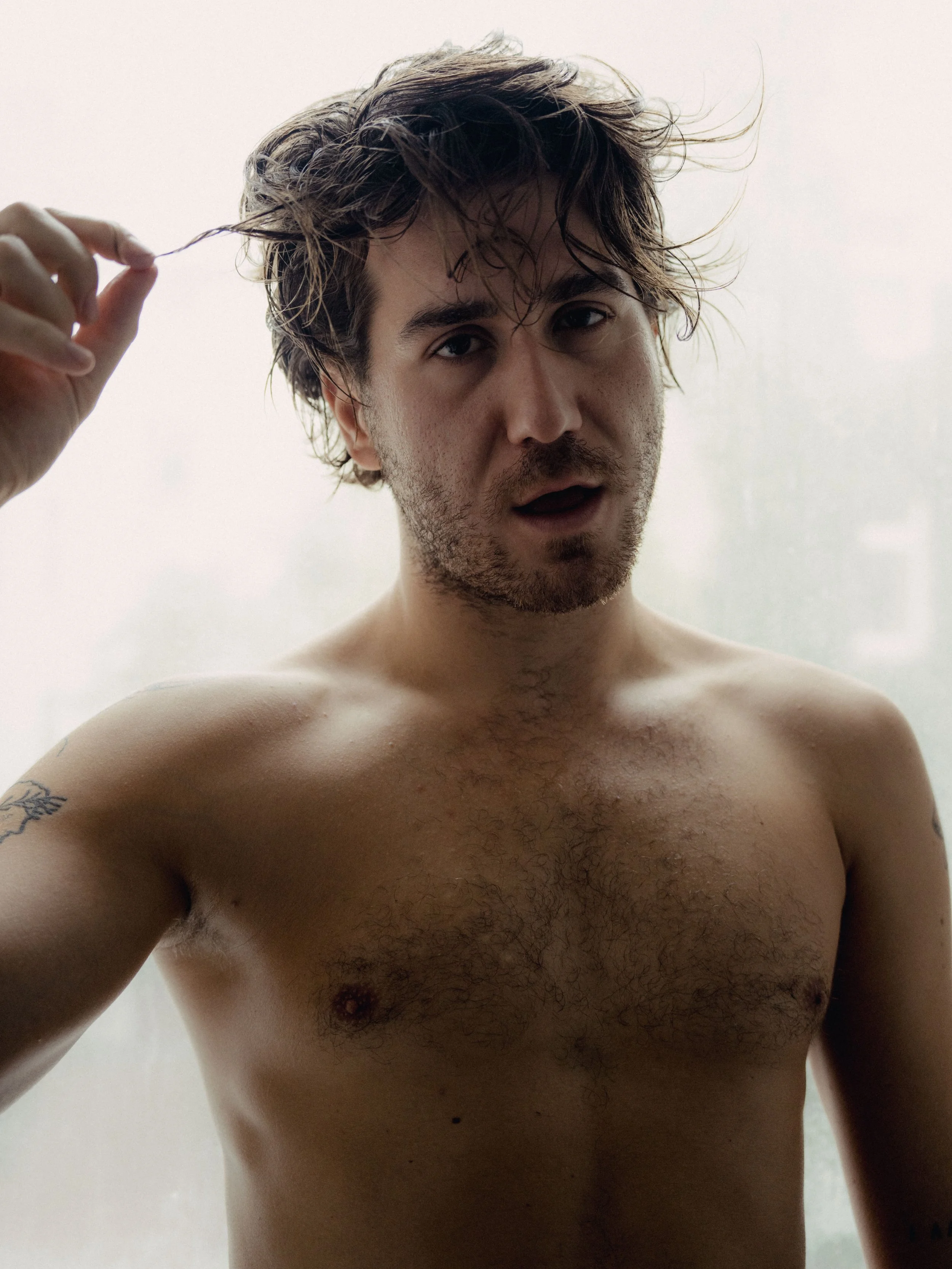 Close-up of a shirtless young man with messy hair, looking at the camera, raising his hand to his hair.