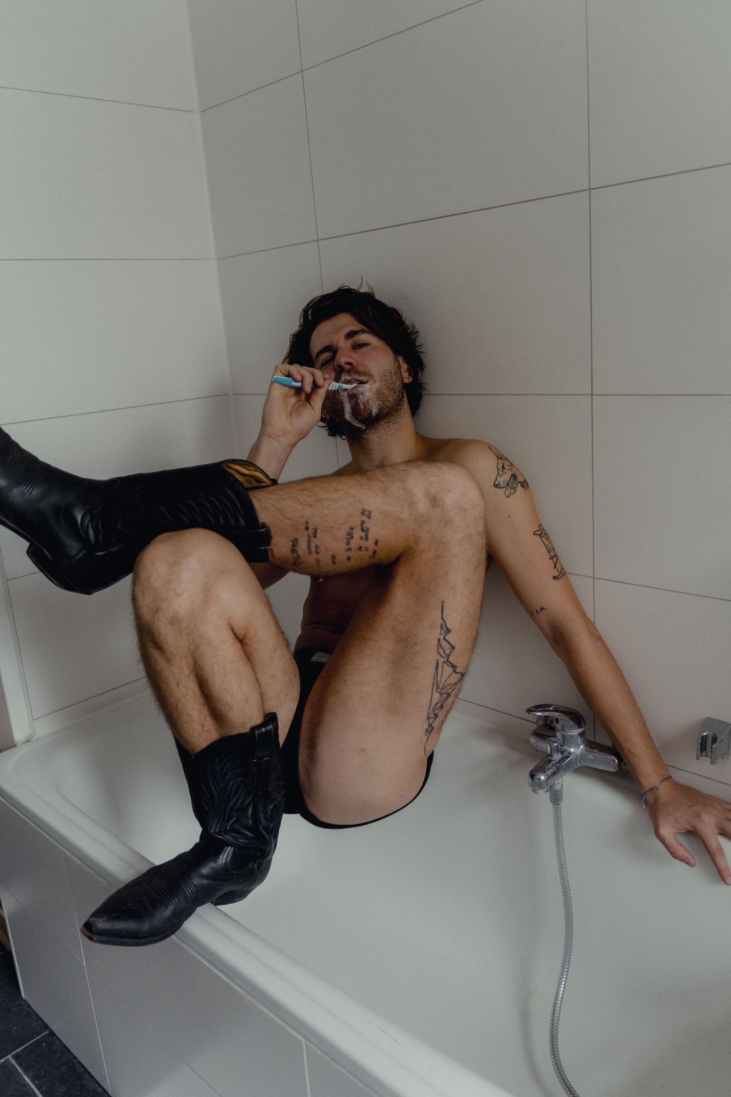 A man with tattoos and black boots sitting naked in a bathtub, brushing his teeth, with a foot in a boot touching his face.