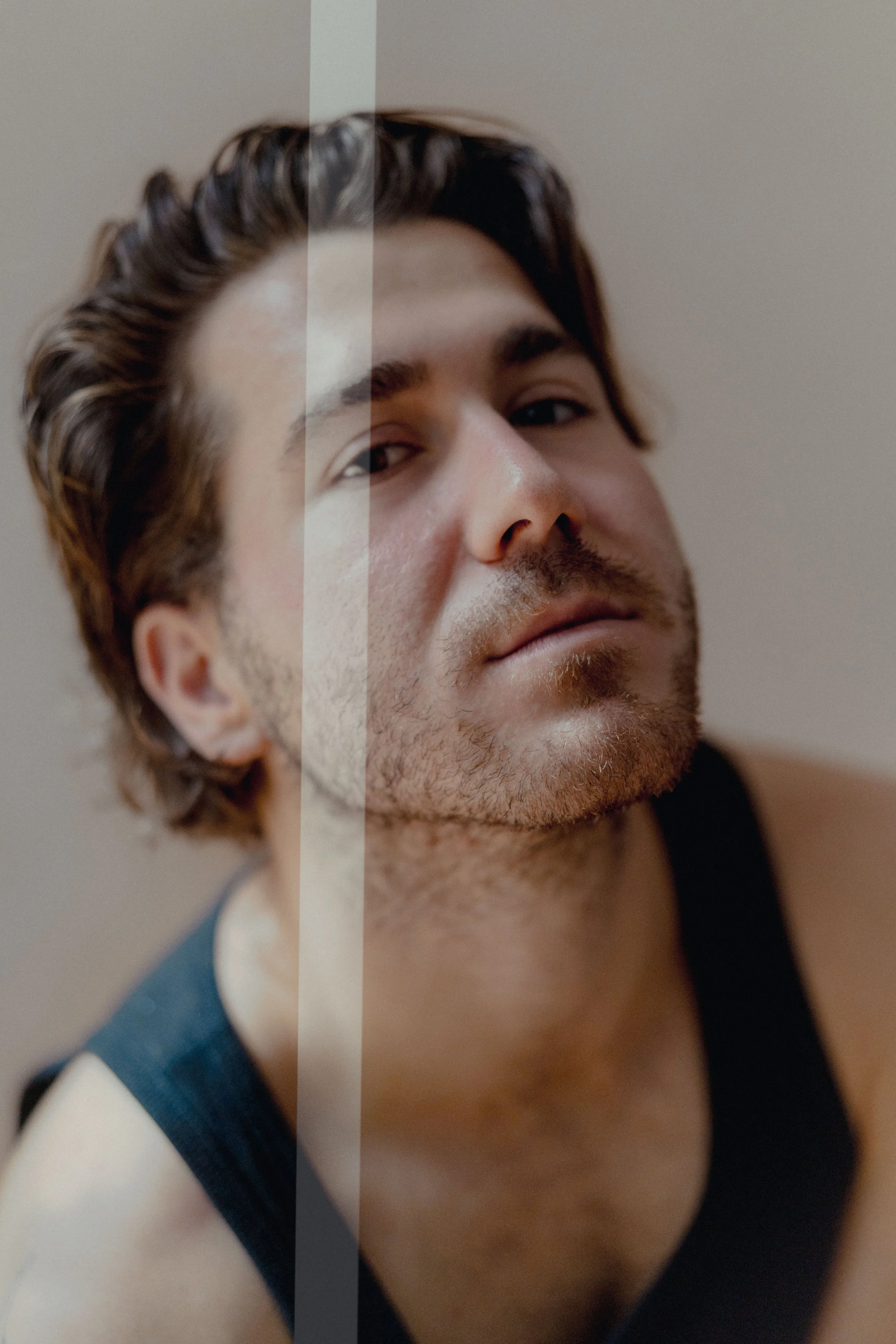 Close-up of a young man with dark hair, slight beard, and wearing a black sleeveless shirt, looking at the camera.