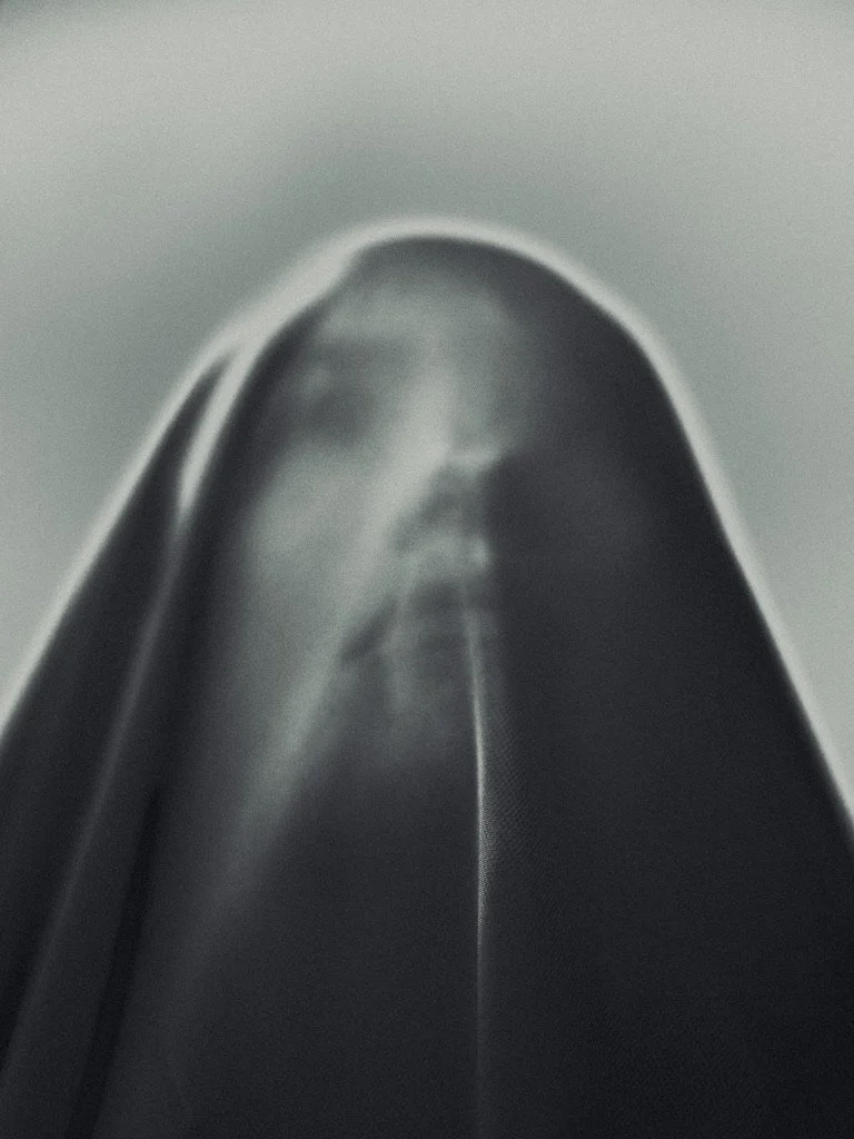 A person in a dark hooded garment, possibly a cloak or hoodie, looking down with their face partially visible in shadow.