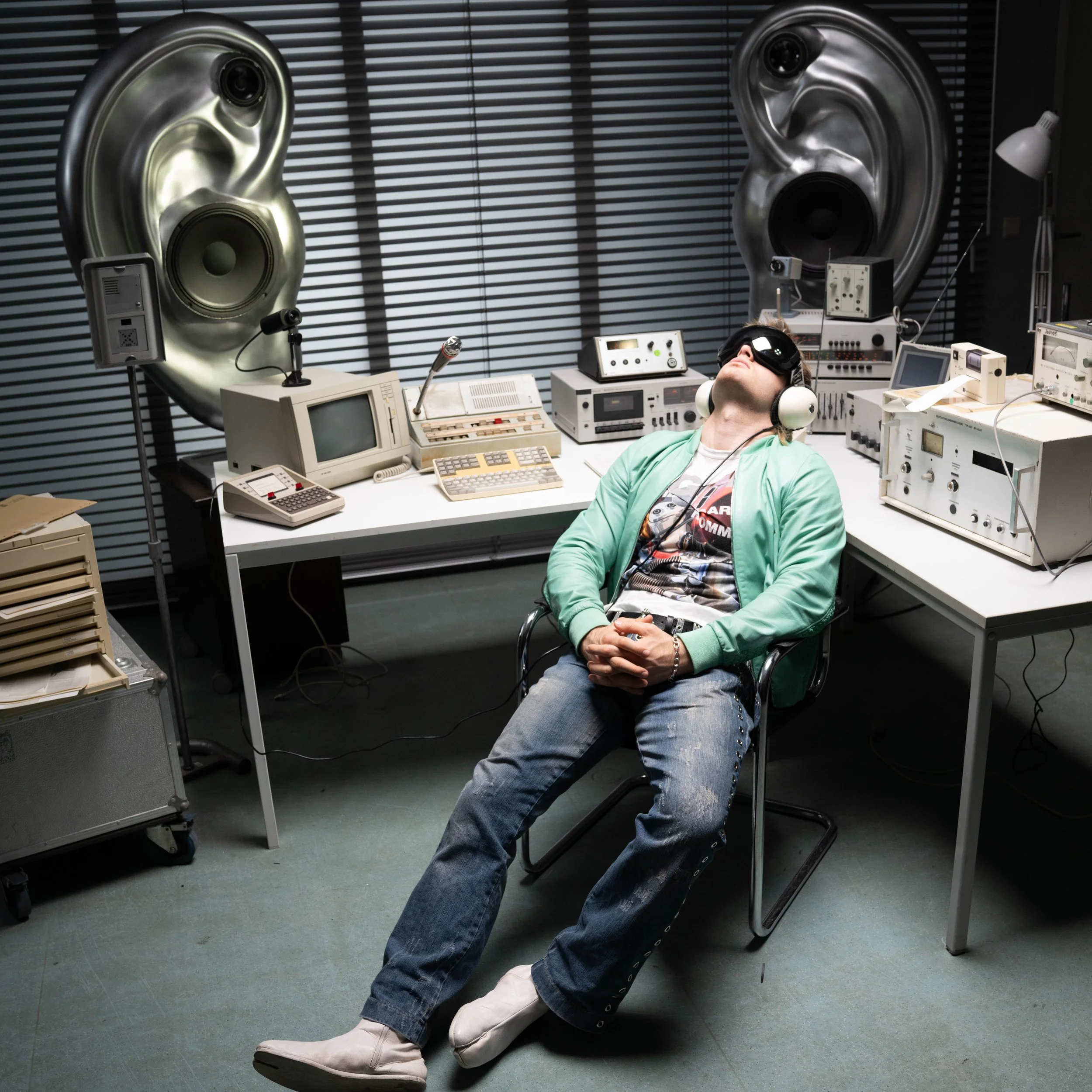 A person with a virtual reality headset and headphones reclining in a cluttered room filled with vintage computer and audio equipment, with large metallic ear sculptures on the wall behind them.