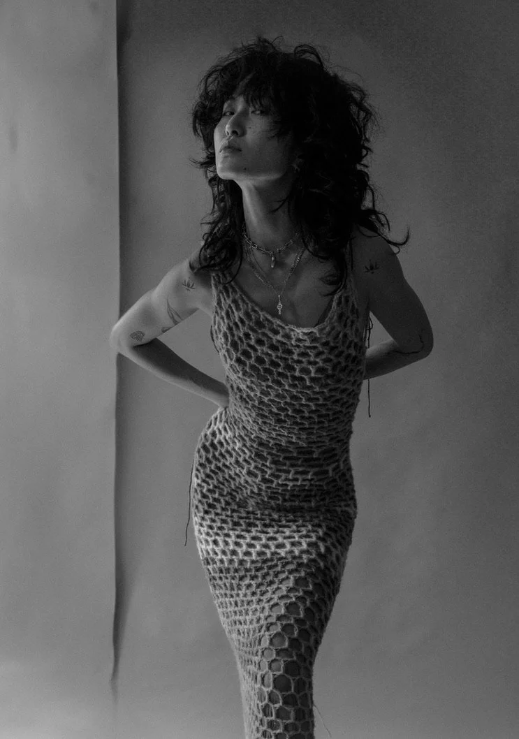 A woman with curly hair wearing a patterned dress, standing with her hands behind her back, looking slightly upwards in a black-and-white photo.