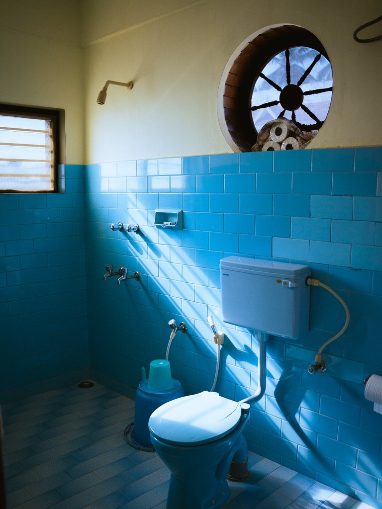 Small bathroom with blue tiled walls, a toilet, a small window, and a circular window with wooden frame.