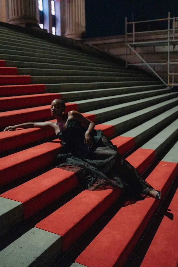 A woman in a black dress lying on red-carpeted steps at night with a building and scaffolding in the background.