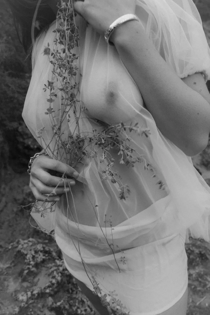 A person holding some wildflowers, wearing a sheer, flowing fabric with their arm and part of their torso visible, outdoors.