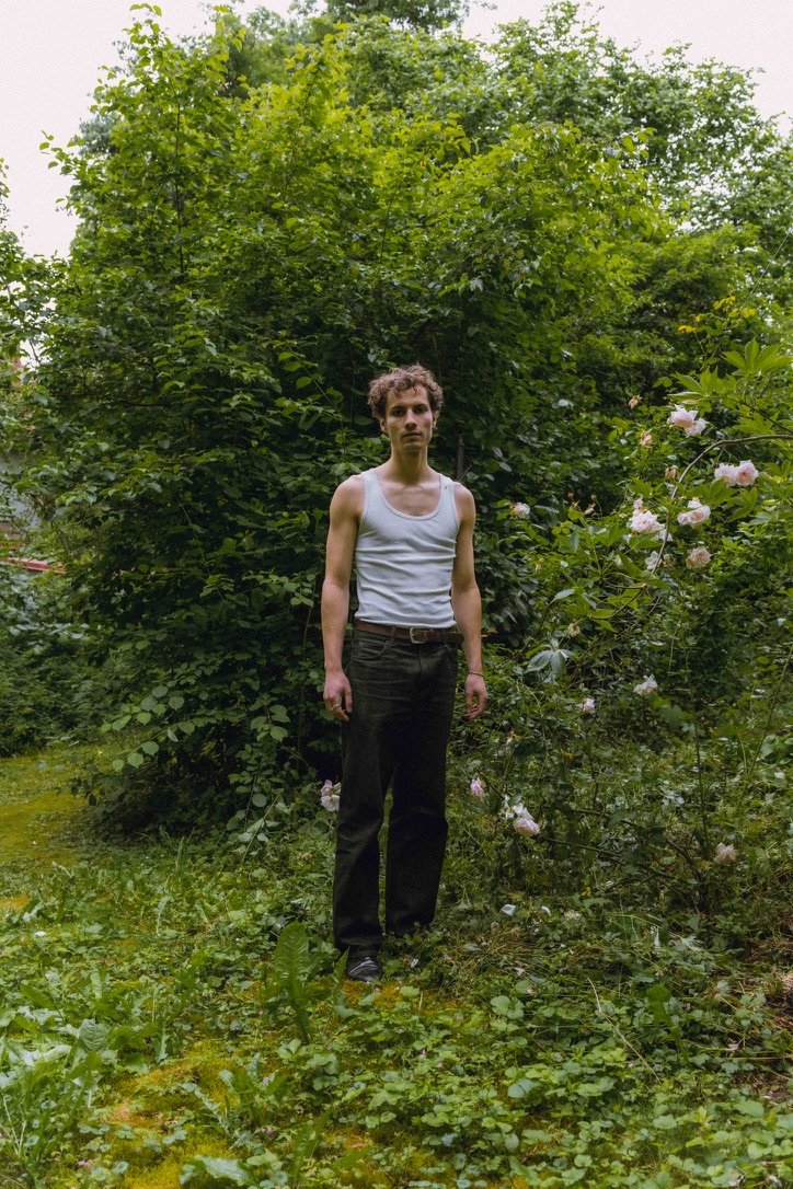 A young man with curly hair wearing a white tank top and black pants standing outdoors in front of lush green bushes and trees, with pink flowers on the right side.