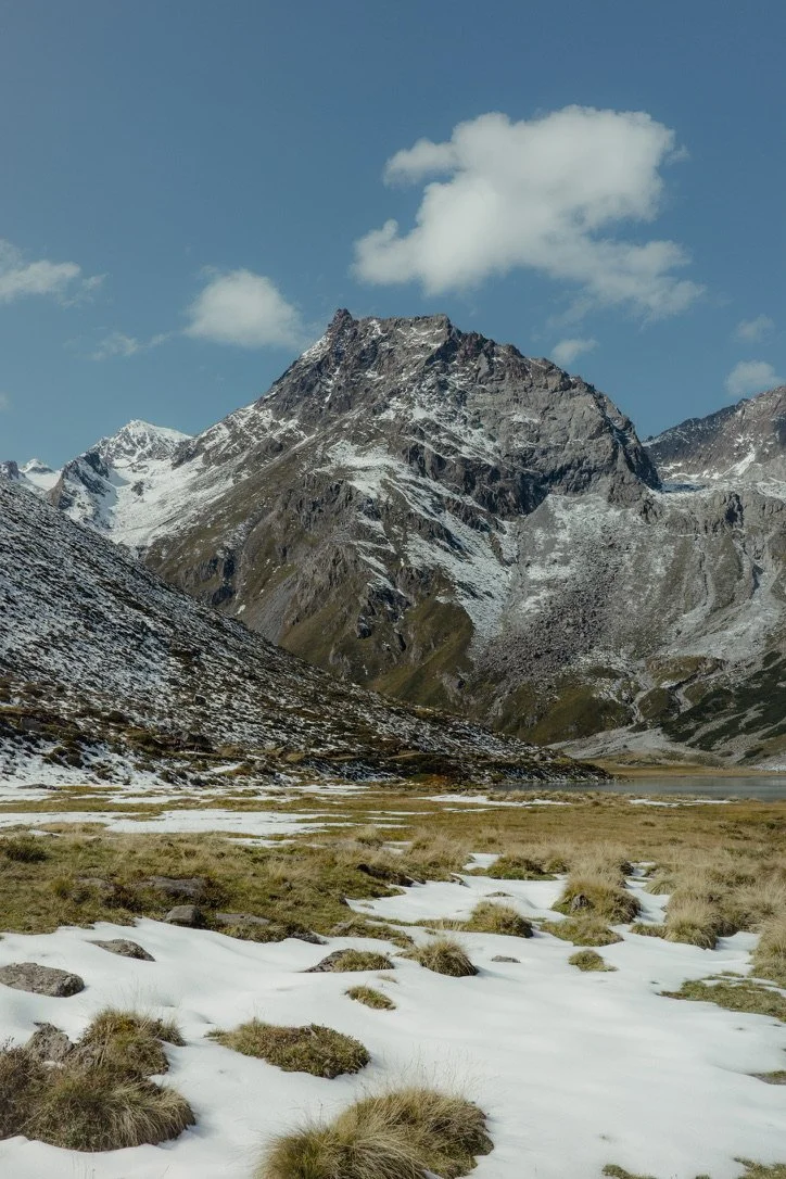 A mountain landscape with snow on the ground and on the mountain slopes, grassy patches in the foreground, and a partly cloudy sky.