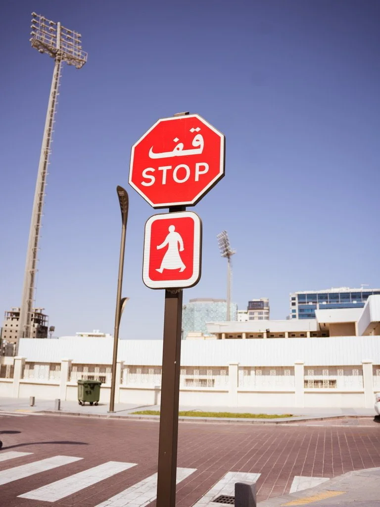 A stop sign with Arabic and English text, and a pedestrian symbol, located on a city street near tall buildings and stadium lights.