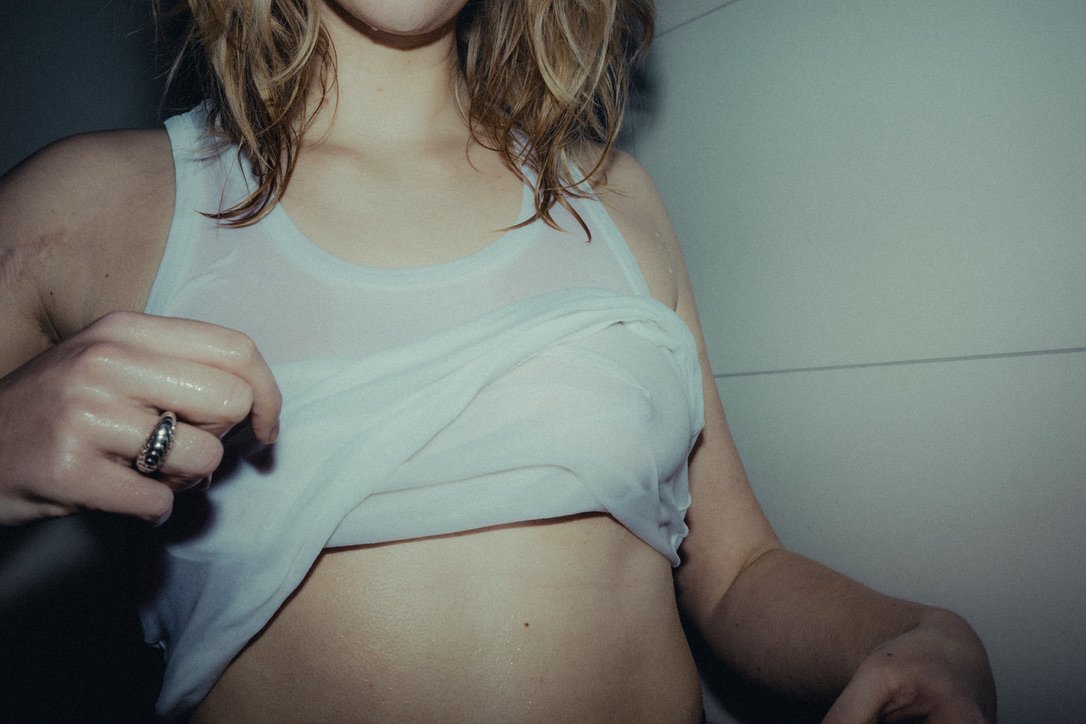 Close-up of a woman wearing a white sleeveless top, lifting it to reveal her bare midriff, with her hand displaying a ring.