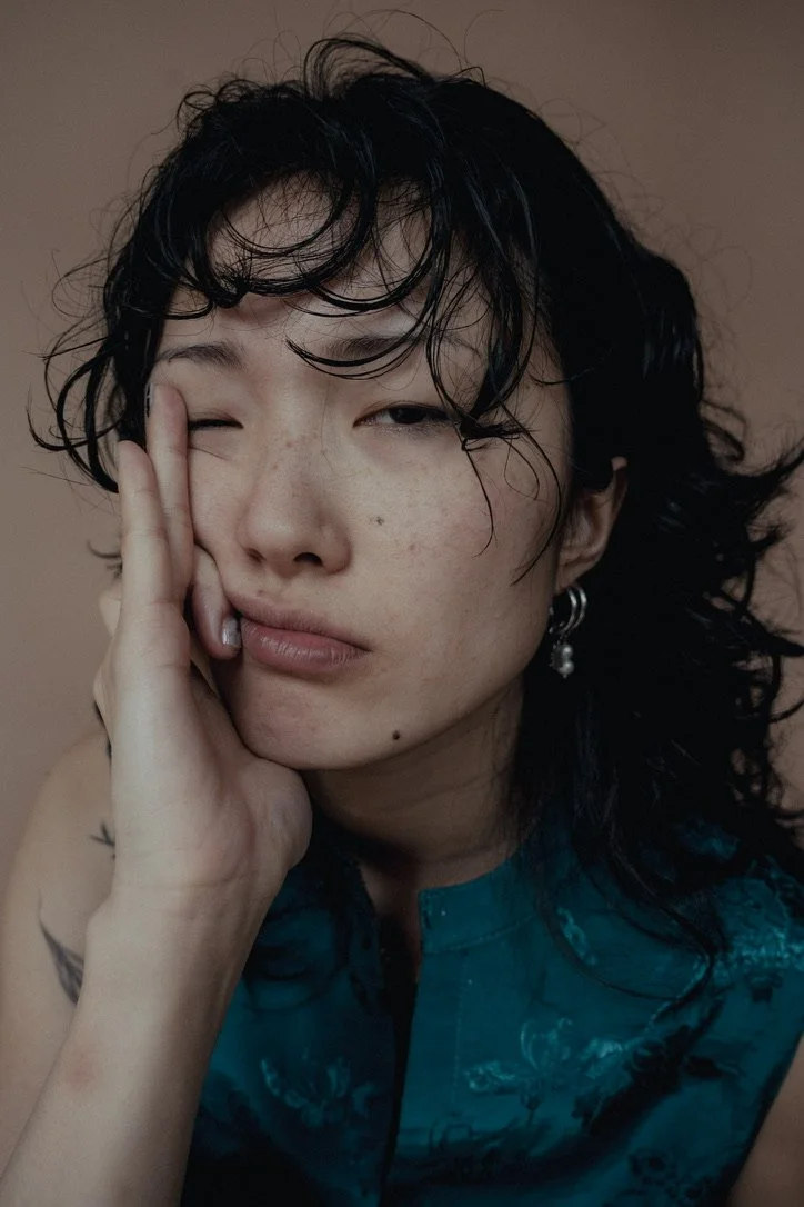 A person with dark, curly hair and light skin, resting their face on their hand, winking at the camera. They are wearing a blue patterned top and earrings.