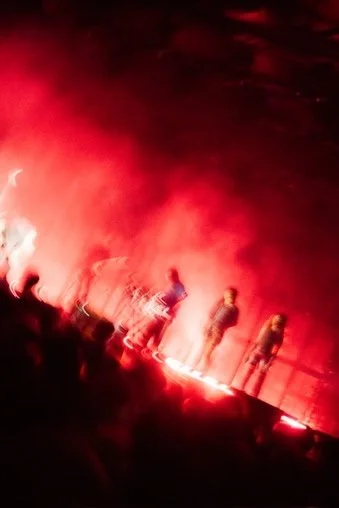 People standing on a bridge with red flares illuminating the scene at night.
