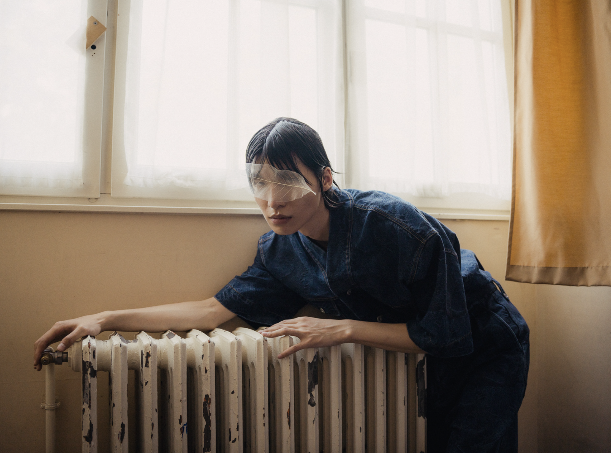 A woman with short dark hair wearing glasses and a denim jacket is stretching or reaching over a radiator, with a window in the background and a yellow curtain to the right.