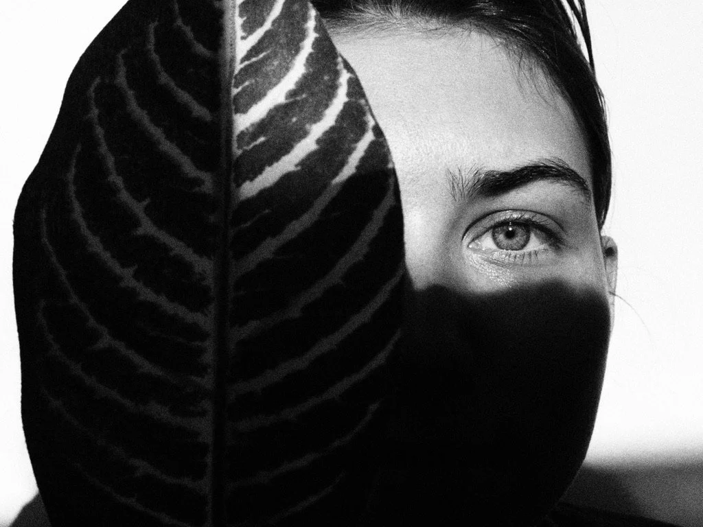 A person partially obscured by a large striped leaf, with only one eye and part of the face visible in black and white.