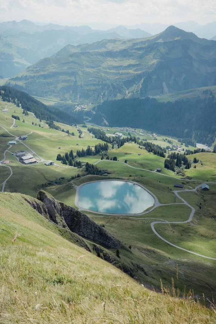 A scenic mountain landscape with rolling green hills, a small lake reflecting the sky, and distant mountain ranges under a partly cloudy sky.