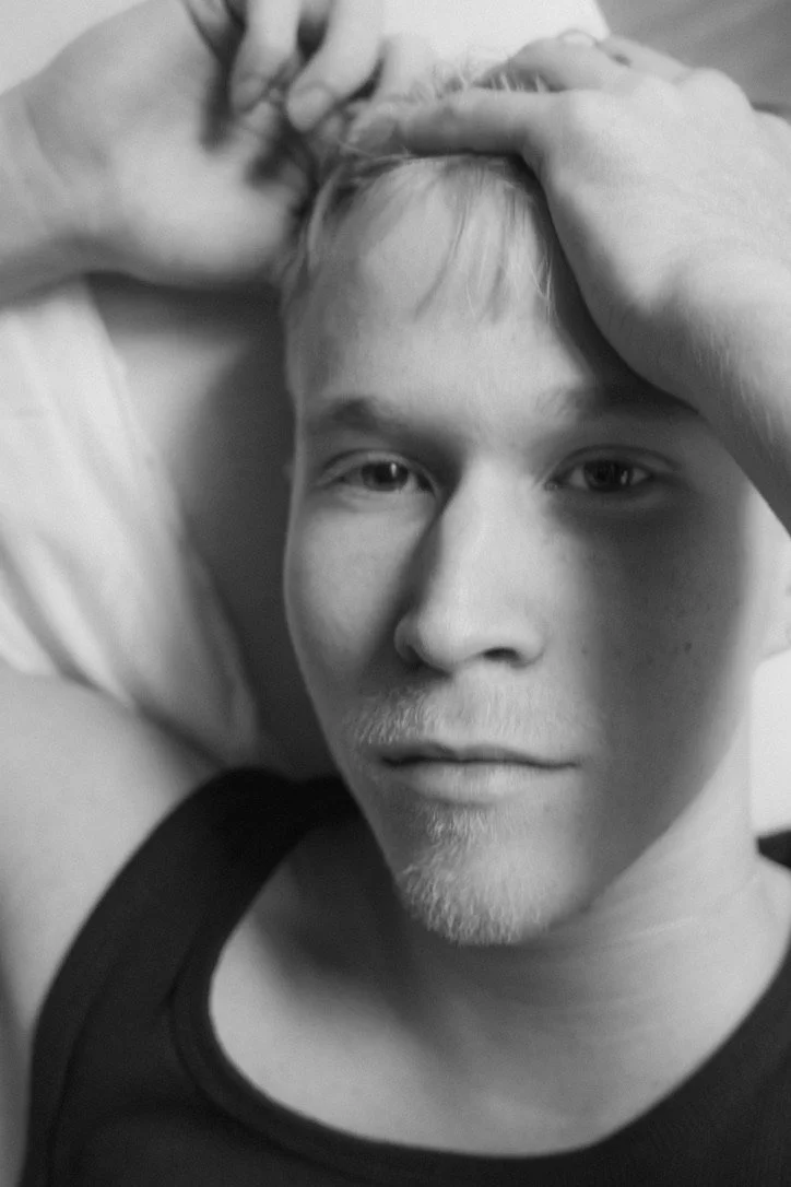 A black-and-white close-up photo of a young man lying on a pillow, with one hand resting on his forehead, looking directly at the camera.