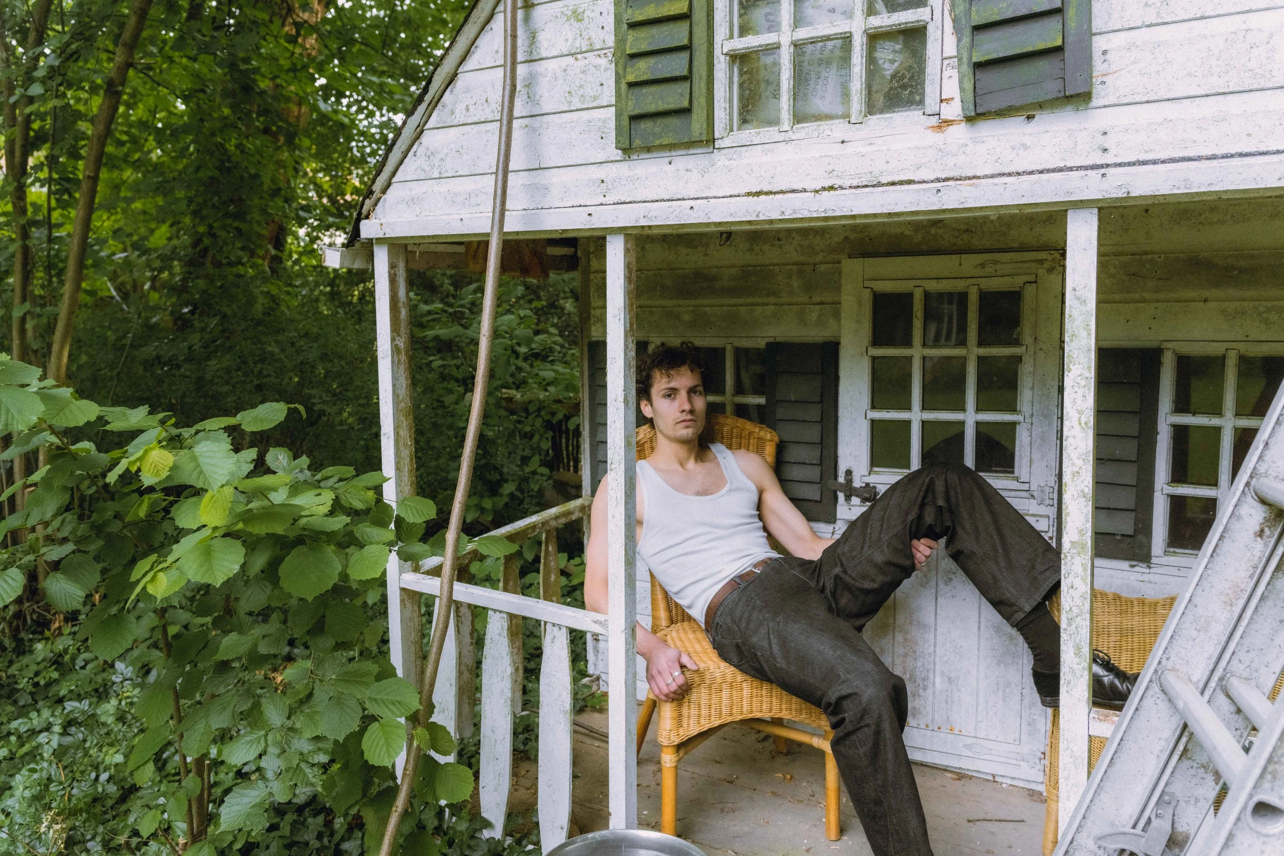 A young man with short curly hair, wearing a light-colored tank top and dark pants, is sitting on a wicker chair on the porch of a weathered, white wooden house with green shutters, surrounded by lush green foliage.