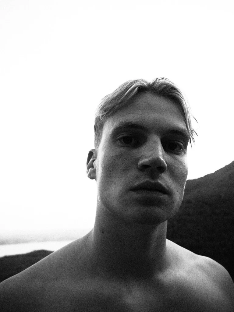 A black and white close-up portrait of a young man with light hair, facing the camera, outdoors with a mountain and cloudy sky in the background.