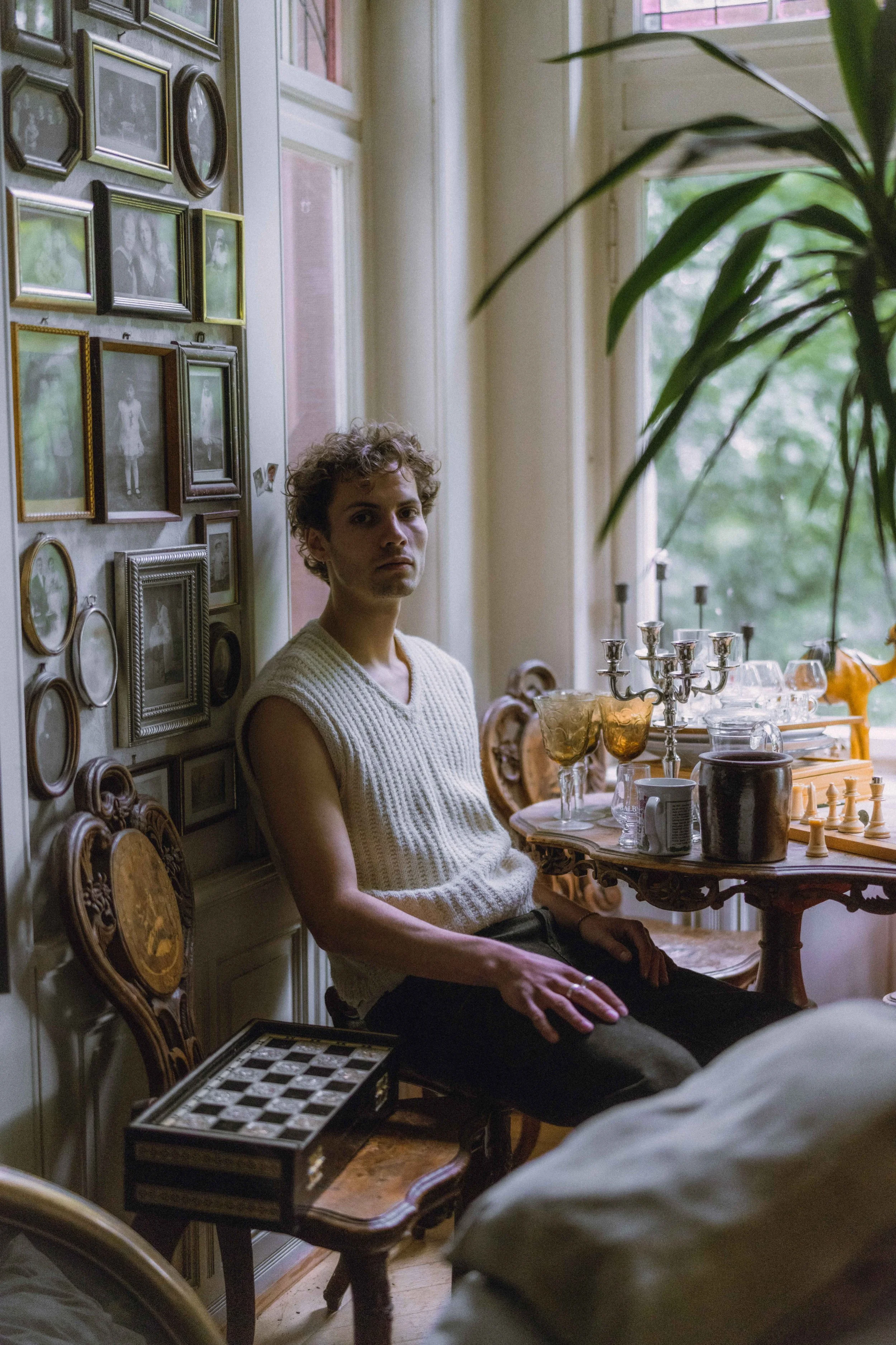 A young man with curly hair sitting in a vintage-style room with a wall of framed photos, a chessboard on a small table, and a window with greenery outside.