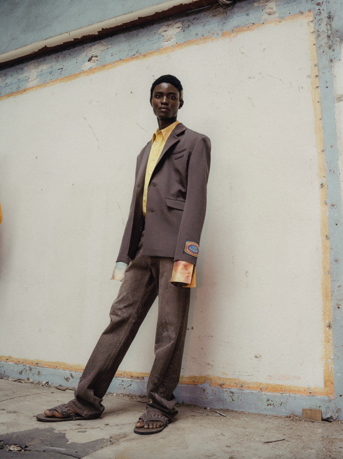 Fashionably dressed young man standing against a weathered wall, wearing a brown suit jacket over a yellow shirt, brown pants, and sandals.
