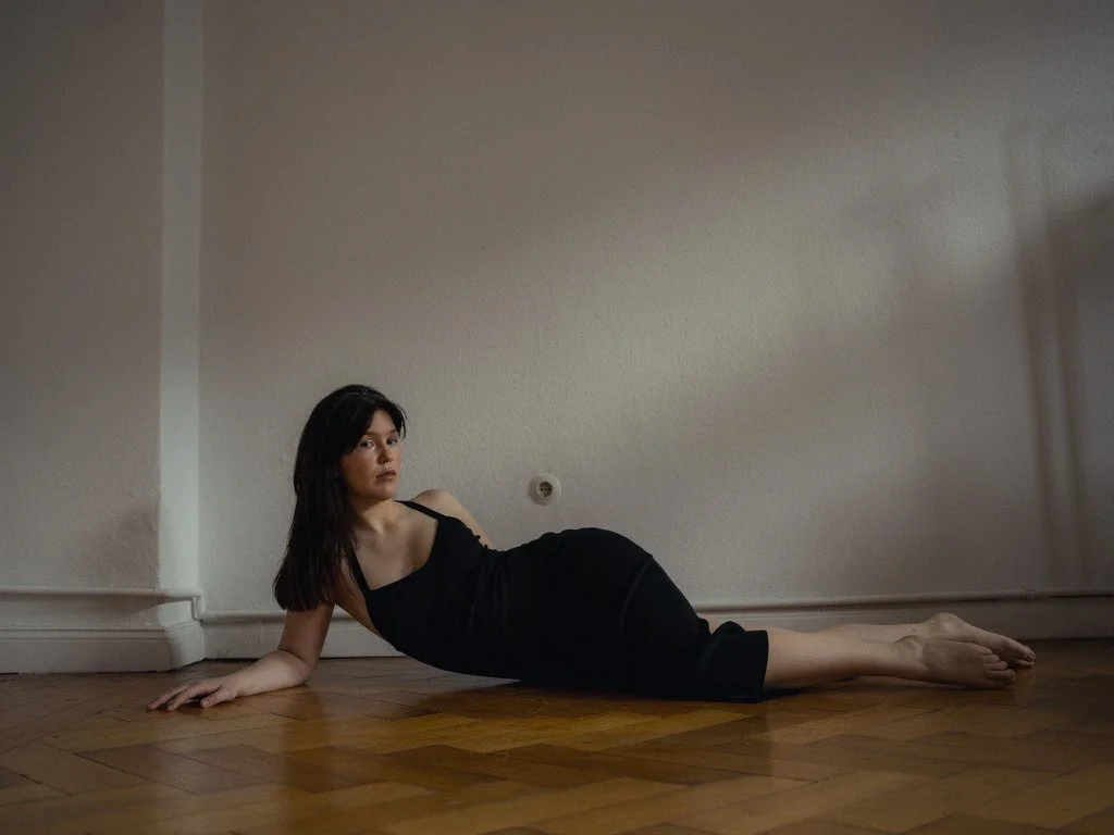A woman in a black dress is lying on a wooden floor, propped up on her left arm with her legs extended to the right, in a minimalist room with plain walls.