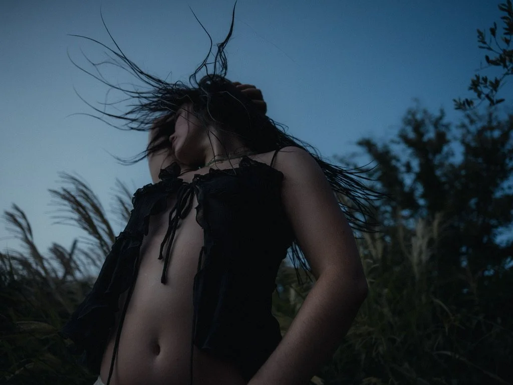 Woman with long hair in motion, wearing a black sleeveless top, outdoors at dusk or dawn, surrounded by tall grasses and trees.