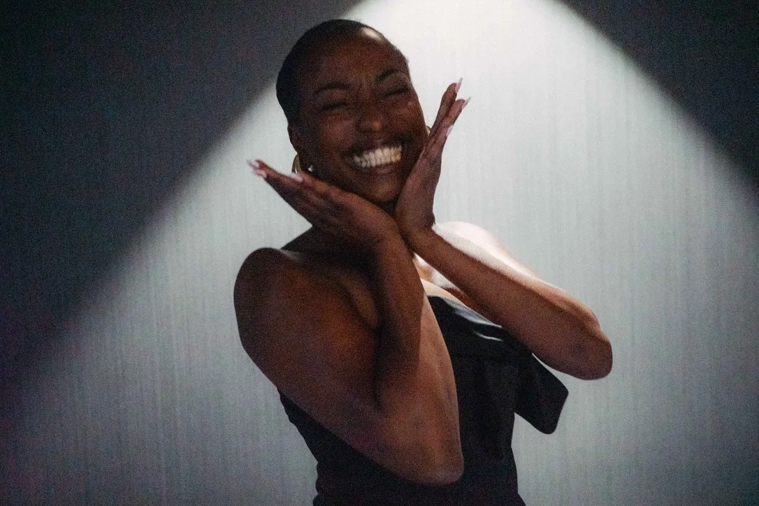 Smiling woman with hands on face, in a dark room with focused lighting.