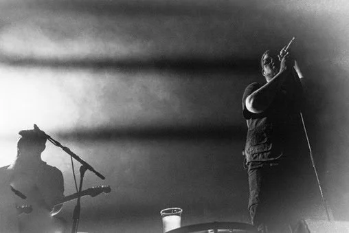 A black and white photo of two musicians performing on stage. One is playing a guitar and sitting down, and the other is singing into a microphone, standing.