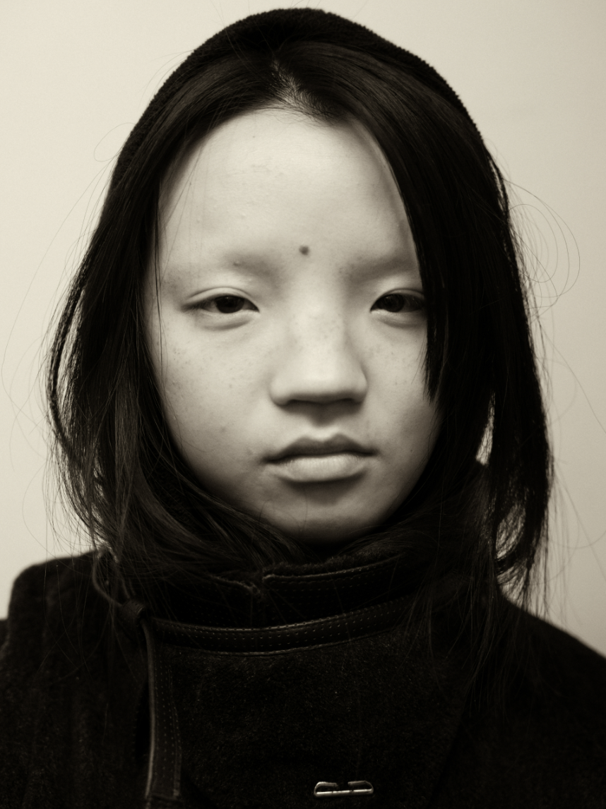 Black and white portrait of a young Asian girl with long, dark hair, wearing a dark jacket, looking directly at the camera with a neutral expression.