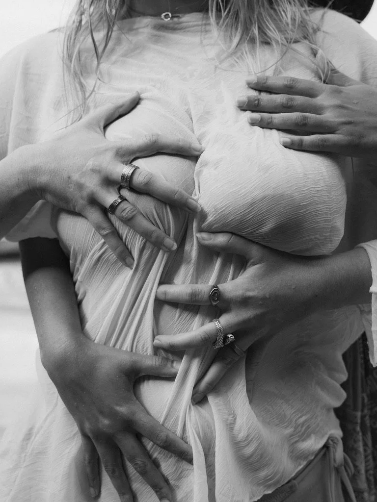 Multiple hands with rings and jewelry form a heart shape on a person's large pregnant belly.