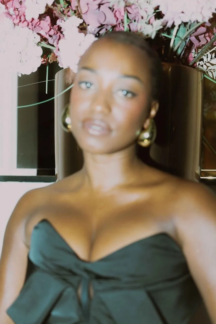 A woman lying down on a surface surrounded by pink flowers, wearing a black strapless dress and large gold hoop earrings.
