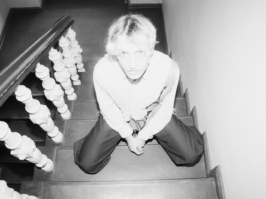 A person with light-colored hair sitting on a staircase, looking up at the camera with legs spread and hands clasped between their knees. The staircase has ornate white balusters and a dark handrail.