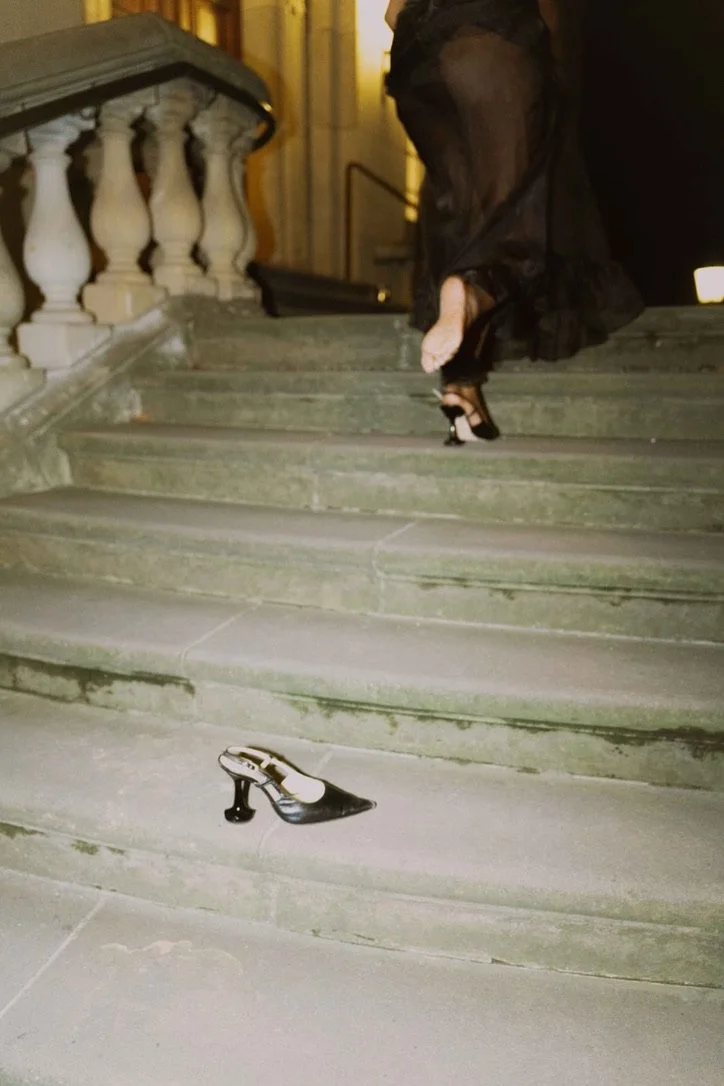 A pair of high-heeled shoes on stone stairs, with a woman holding a high heel in her hand. The shoes are elegant and the stairs are outside a building at night.