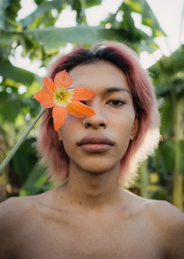 A young woman with pink hair holding an orange flower near her face, surrounded by green foliage.