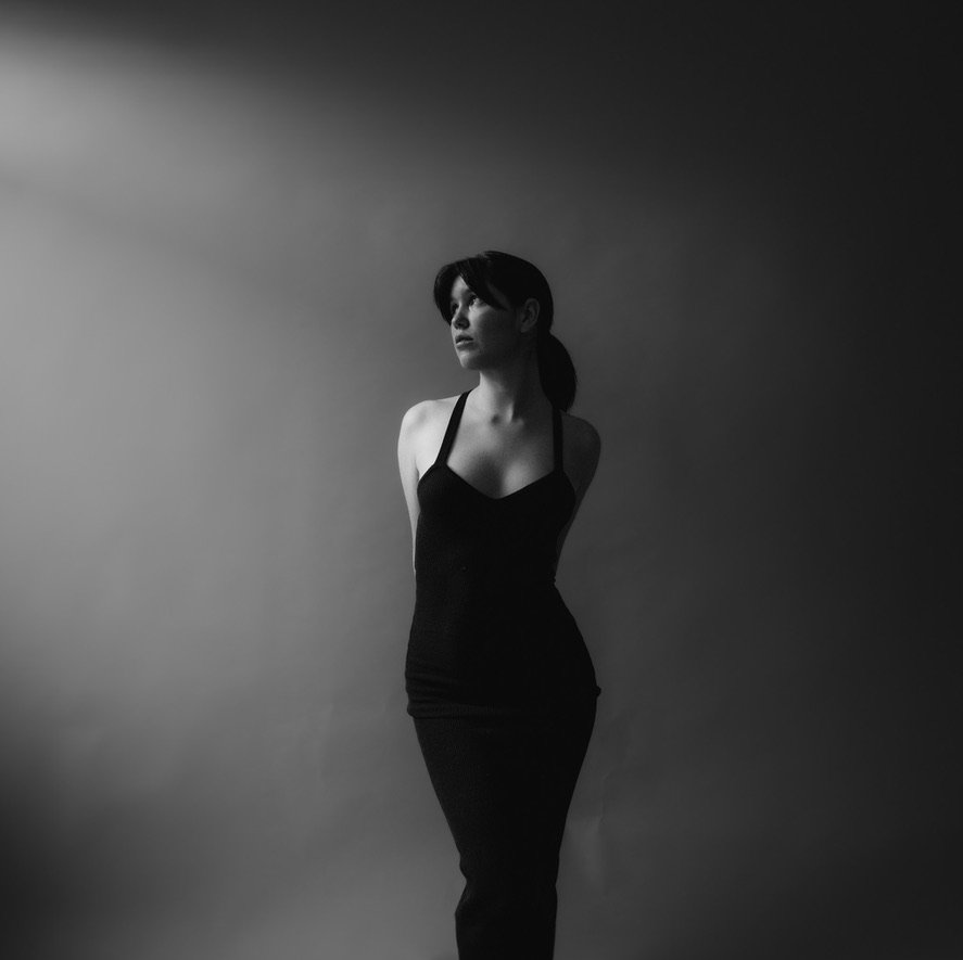 A black and white photo of a woman in a form-fitting black dress, standing against a gradient background with shadows.