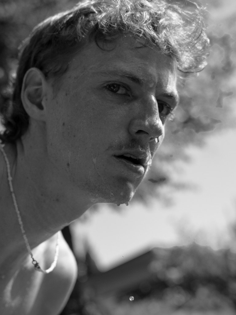A close-up black-and-white photo of a young man with curly hair, looking intently at the camera with a slightly open mouth, outdoors with a blurred background.