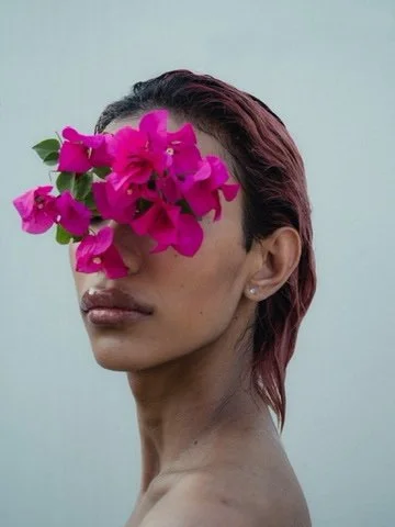 Woman with pink flowers covering her eyes and forehead, with short reddish-brown hair and bare shoulders.
