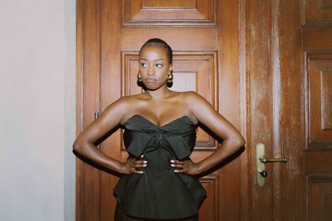 A woman in a strapless black dress standing with hands on her hips in front of a wooden door.