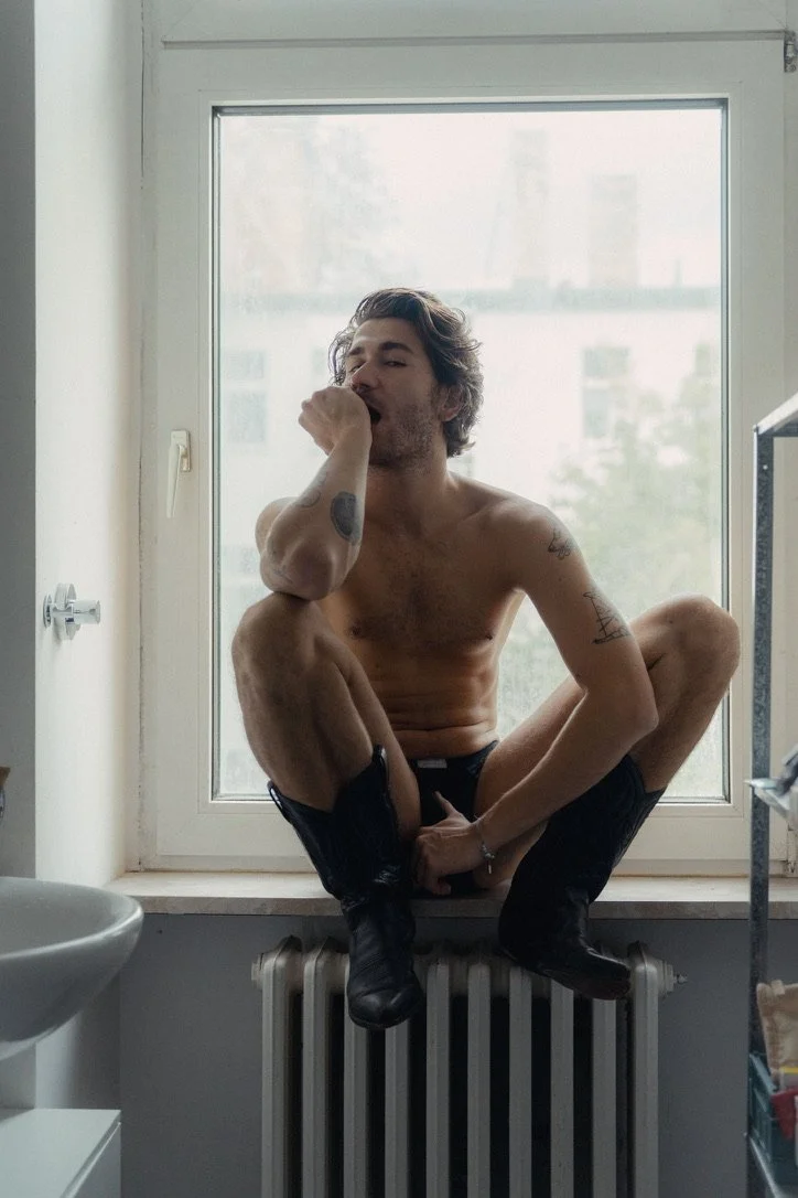 A shirtless man with tattoos sitting cross-legged on a window ledge, wearing black shoes and underwear, with one hand covering his mouth, in front of a large window with a blurred cityscape in the background.