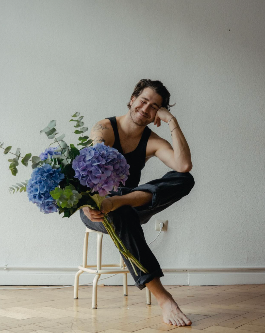 A man sitting on a small stool, holding a bouquet of purple and blue hydrangeas, smiling with eyes closed, wearing a black tank top and black pants, in a minimalist room with a plain white wall and wooden floor.