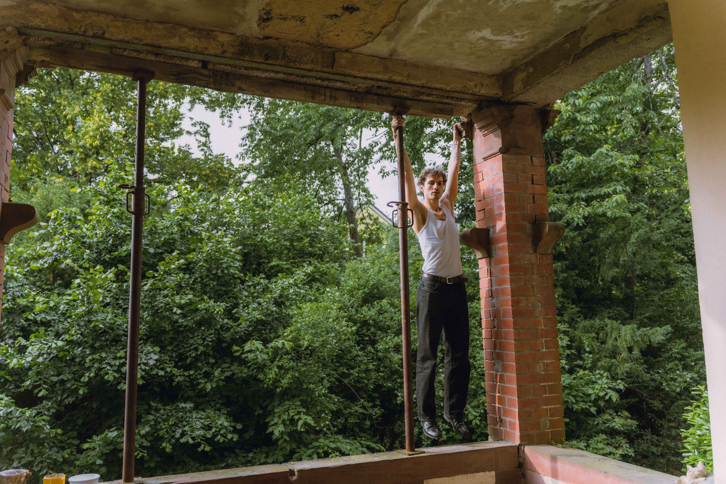 A young man in a white tank top and black pants is holding onto scaffolding poles with both hands, standing on a partially built porch with a brick column, greenery and trees in the background.