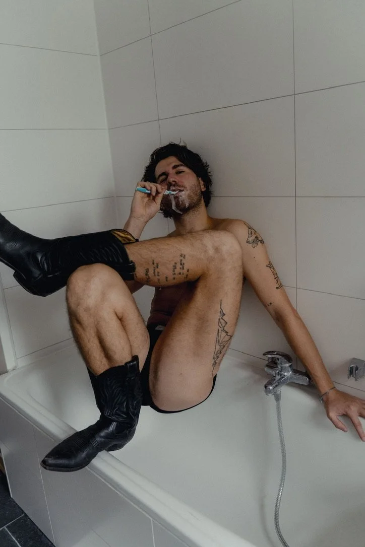 A young man with tattoos sitting in a bathtub, smoking a cigarette, with a person's leg resting on his lap.
