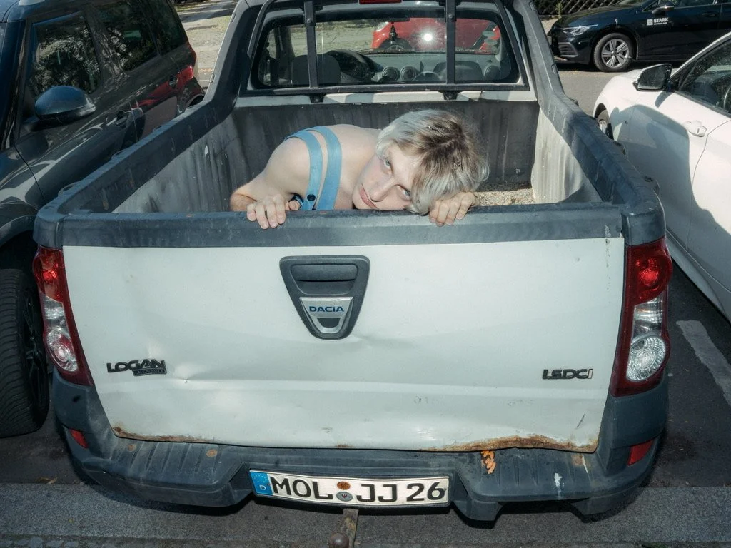 Person with short, light-colored hair lying in the truck bed of a white Dacia Logan pickup truck, peering over the edge and looking at the camera. The truck is parked in a lot with other cars.