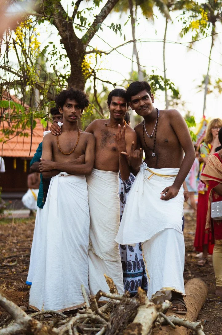 Three men dressed in traditional white dhotis, standing outdoors under a tree. They are shirtless and wearing necklaces. Other people are visible in the background, some dressed in colorful clothing, indicating a cultural or religious gathering.