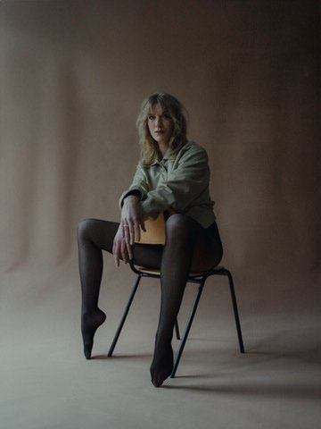 A woman with blonde hair sitting on a black chair, holding a book, wearing a gray jacket and black tights, in a minimalistic studio setting with a plain background.